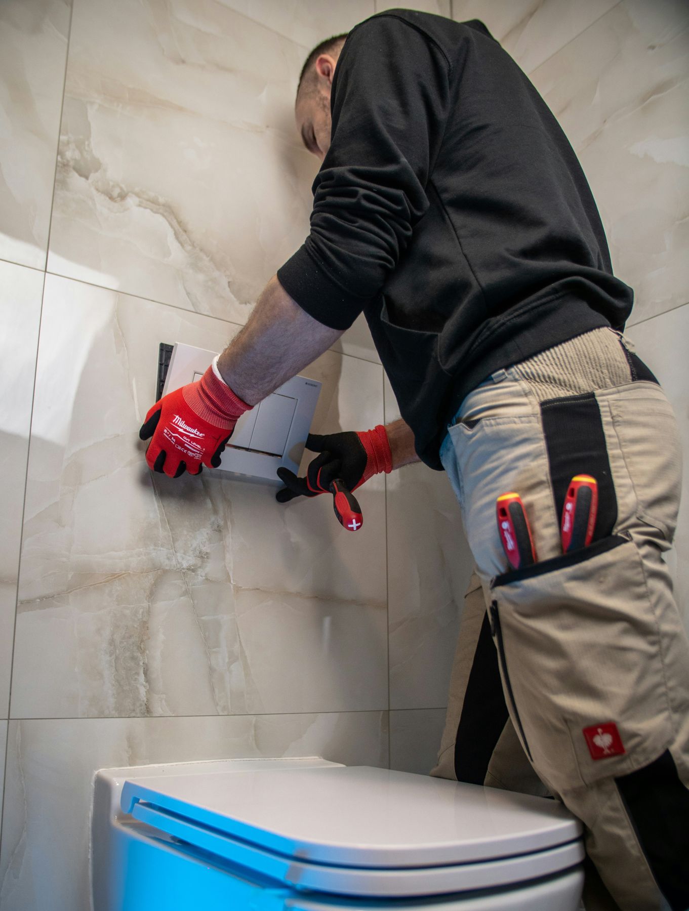 A plumber in work gloves fitting a flush plate on a tiled bathroom wall